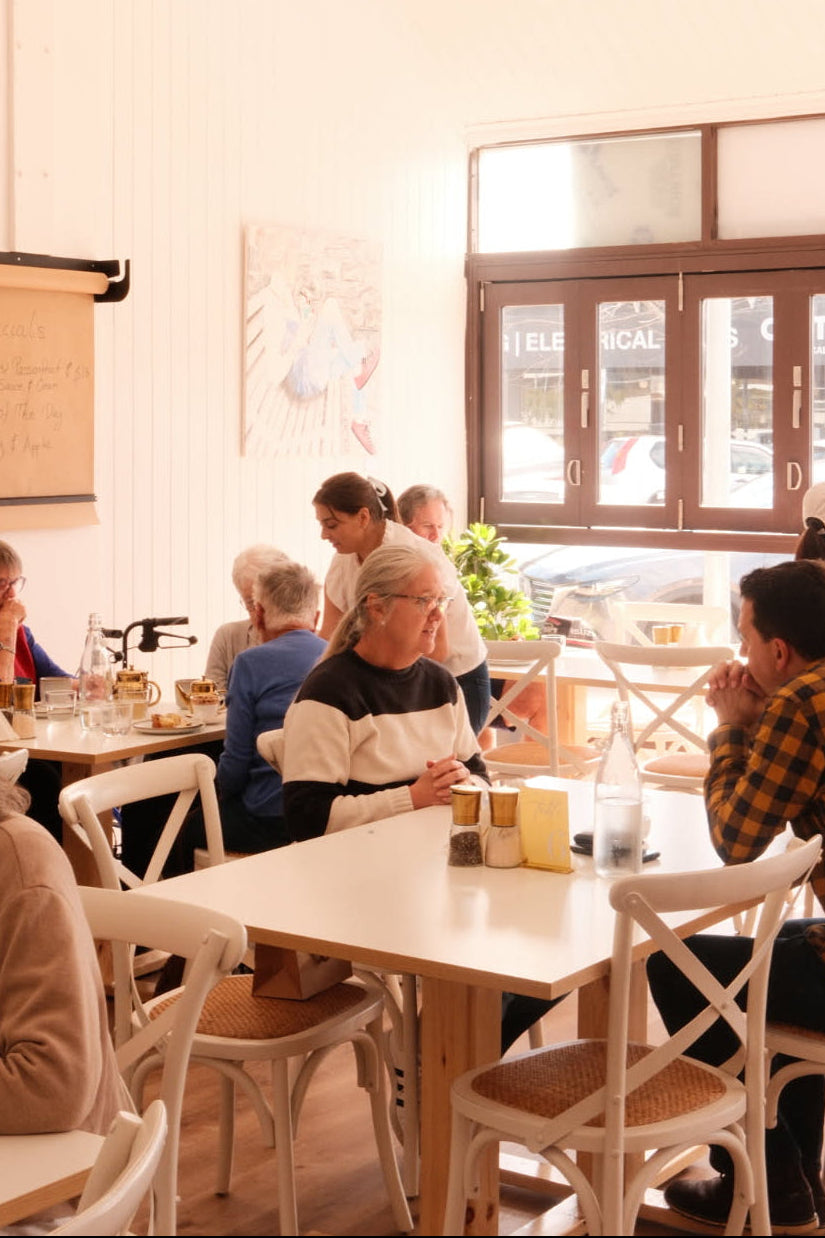 A bright, modern cafe interior with white walls and wooden-framed windows, showing several people seated at white tables with woven-back chairs.