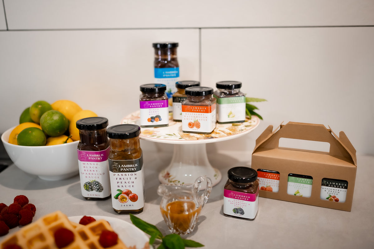 A collection of jarred preserves and spreads from Lambruk Pantry arranged on a white pedestal plate and countertop, alongside a cardboard carrying case. The scene includes fruit, waffles, and a small glass pitcher on a kitchen counter, making this a lifestyle shot.